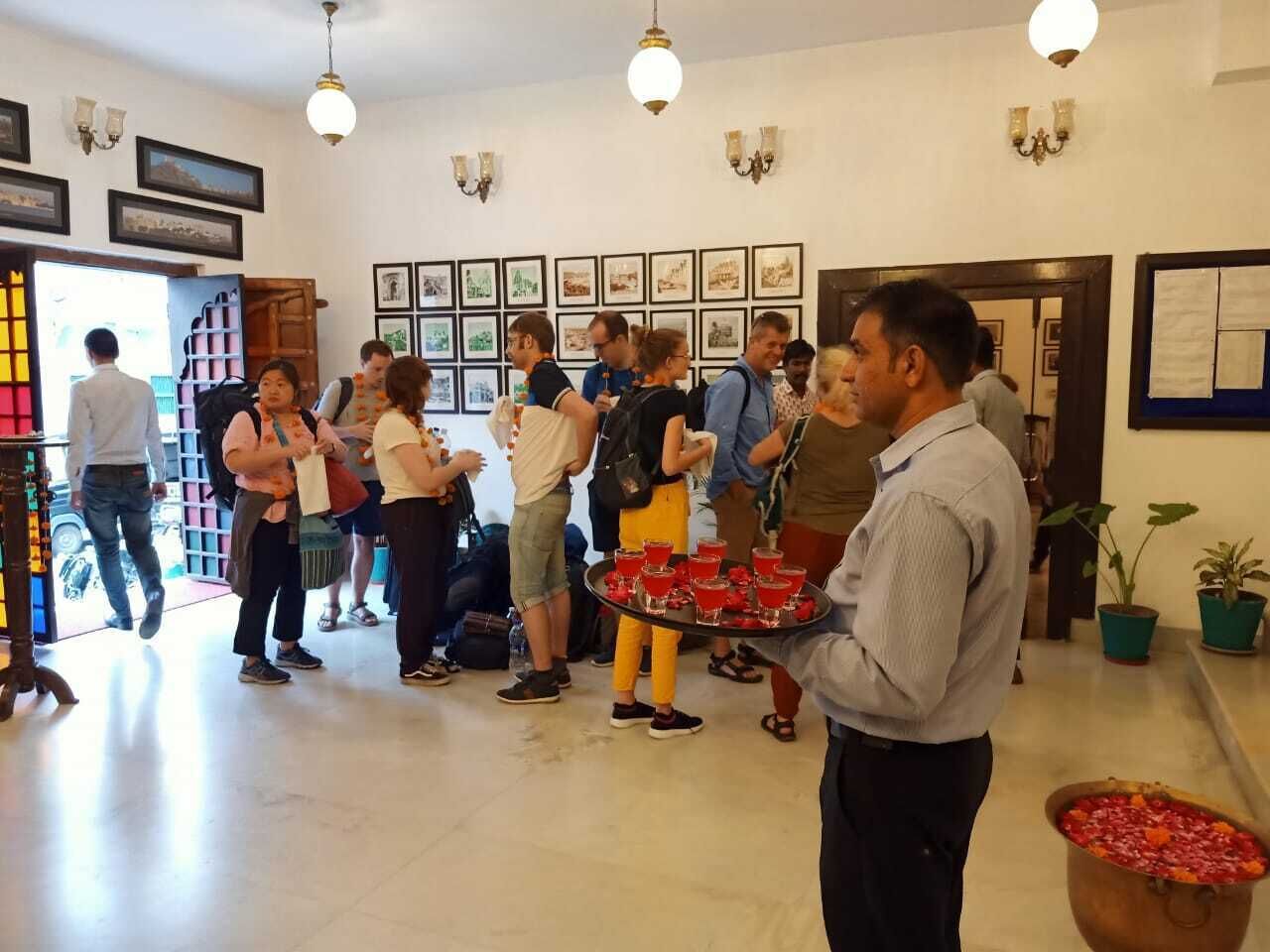 Een hotel lobby met een groep mensen, een man die een dienblad met rode dranken vasthoudt, en een grote pan eten op een tafel.
