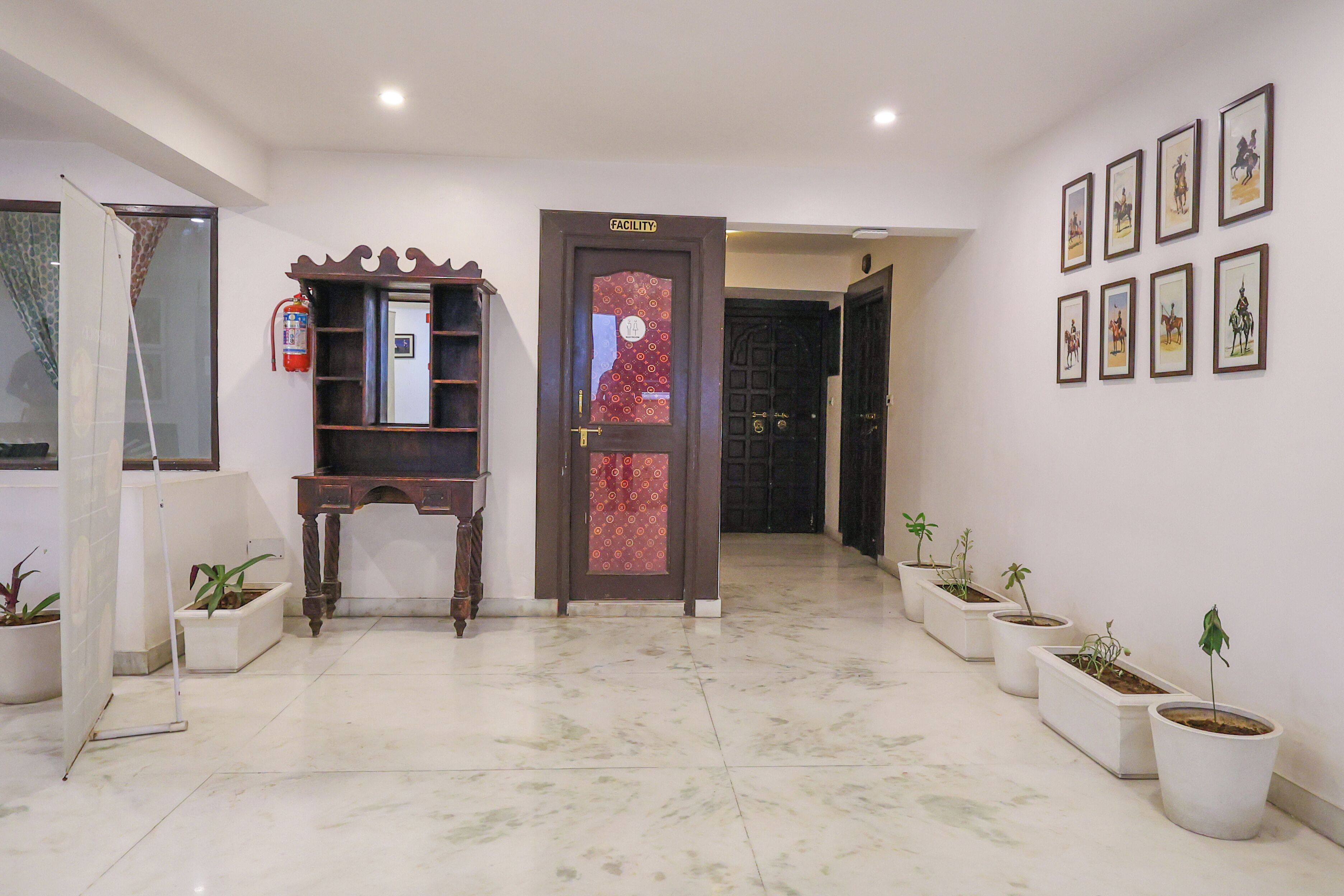 A hallway with a wooden door labeled "FACILITY," potted plants, a fire extinguisher, and framed pictures on the wall.