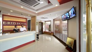 Un espace d’accueil d’hôtel comprenant un comptoir de réception, deux employés, un écran de télévision et un banc.