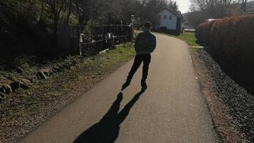 Un chemin pavé sur lequel une personne s’éloigne de la caméra, entourée d’arbres et d’une haie.