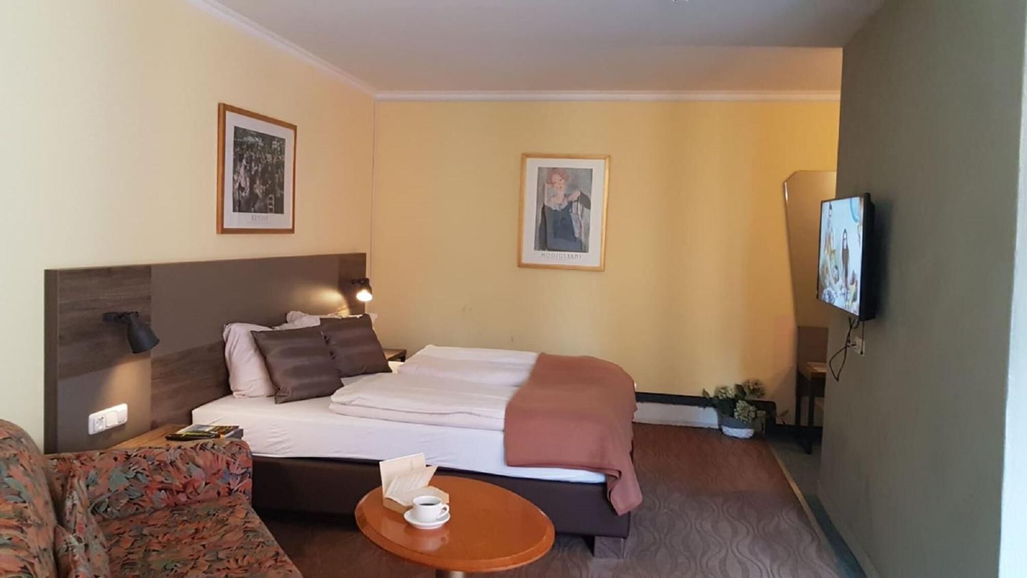 Une chambre d’hôtel avec un lit, un canapé, une petite table avec une tasse, et deux tableaux encadrés au mur.