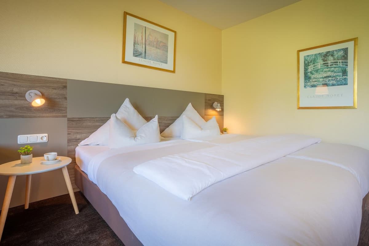 Une chambre d’hôtel avec un grand lit, deux lampes fixées au mur, une petite table avec une plante et deux tableaux encadrés accrochés au mur.
