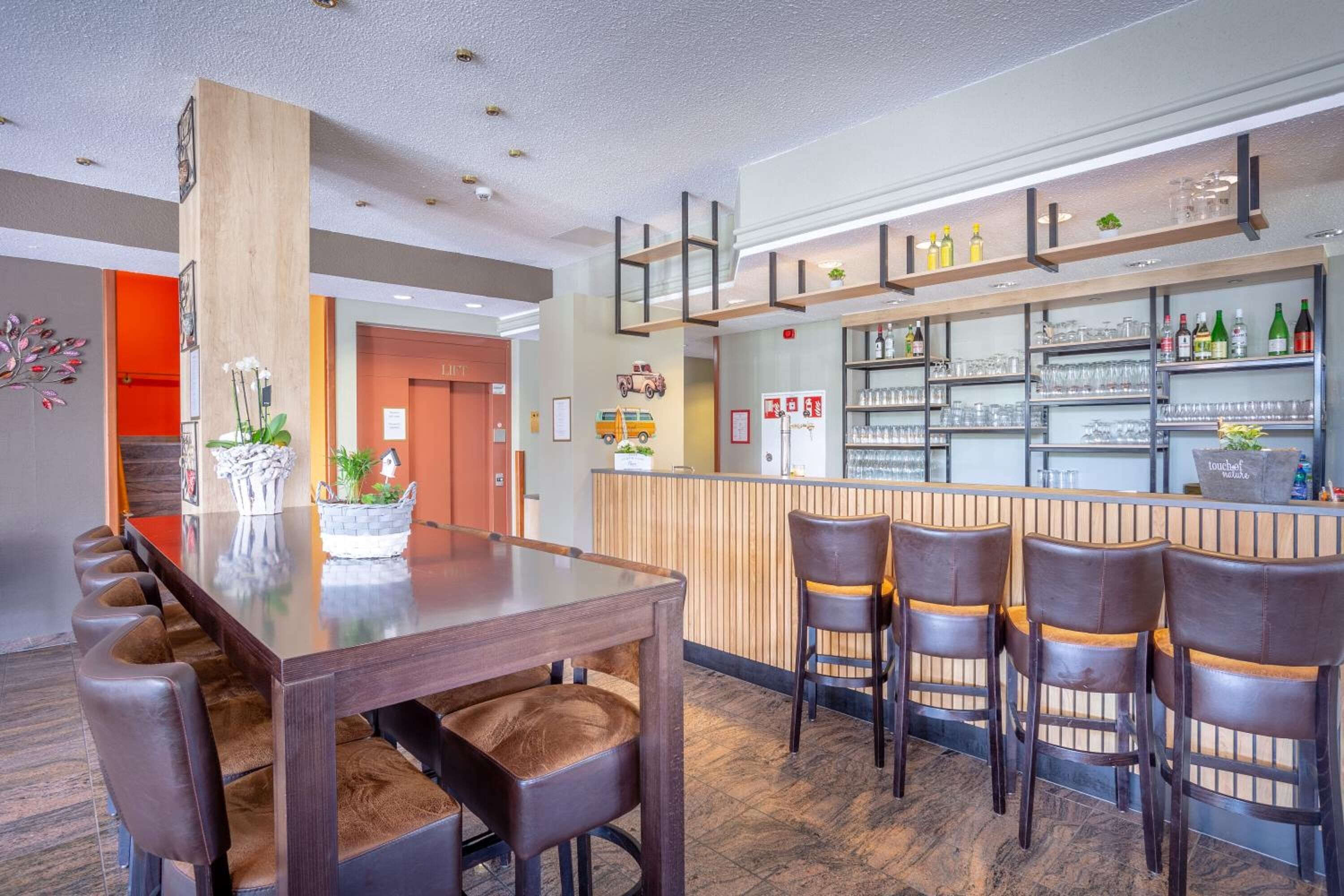 Zona de bar moderna con una larga mesa de madera y sillas de cuero, una barra con estantes llenos de botellas y vasos, y un centro de mesa con planta decorativa.