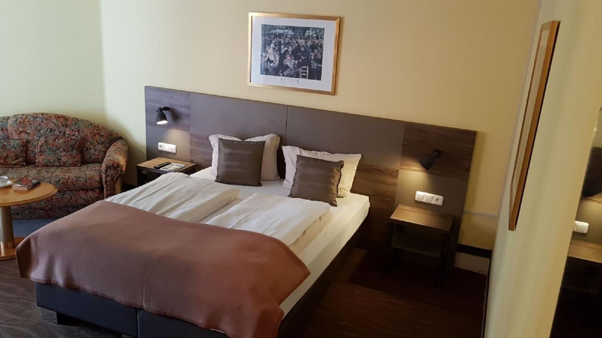 Habitación de hotel con cama, sofá, dos mesitas de noche y un cuadro enmarcado en la pared.