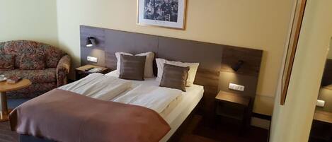 Habitación de hotel con cama, sofá, dos mesitas de noche y un cuadro enmarcado en la pared.