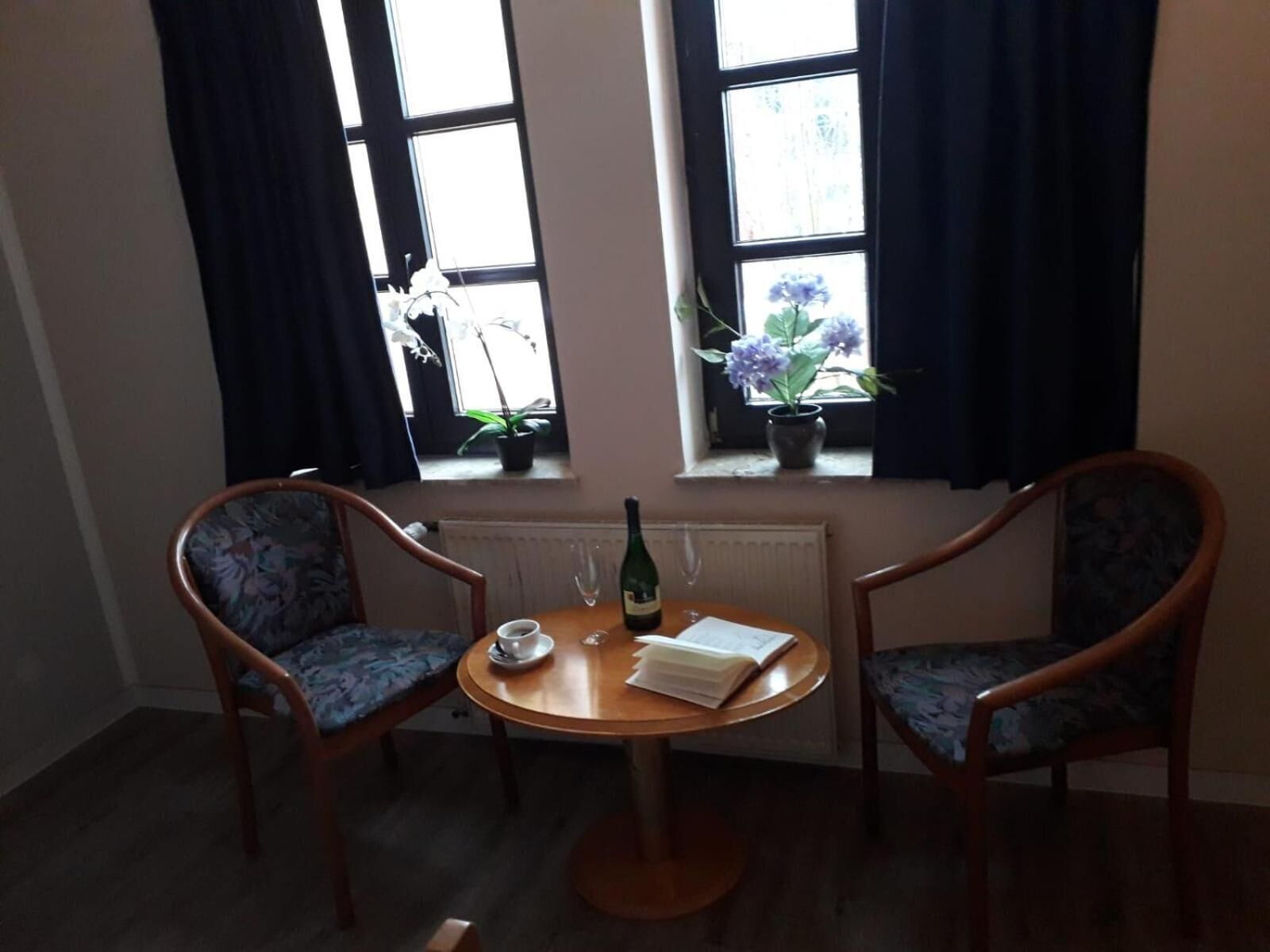 Une petite pièce avec deux chaises en bois, une table ronde, un livre, une bouteille et deux plantes en pot près des fenêtres.