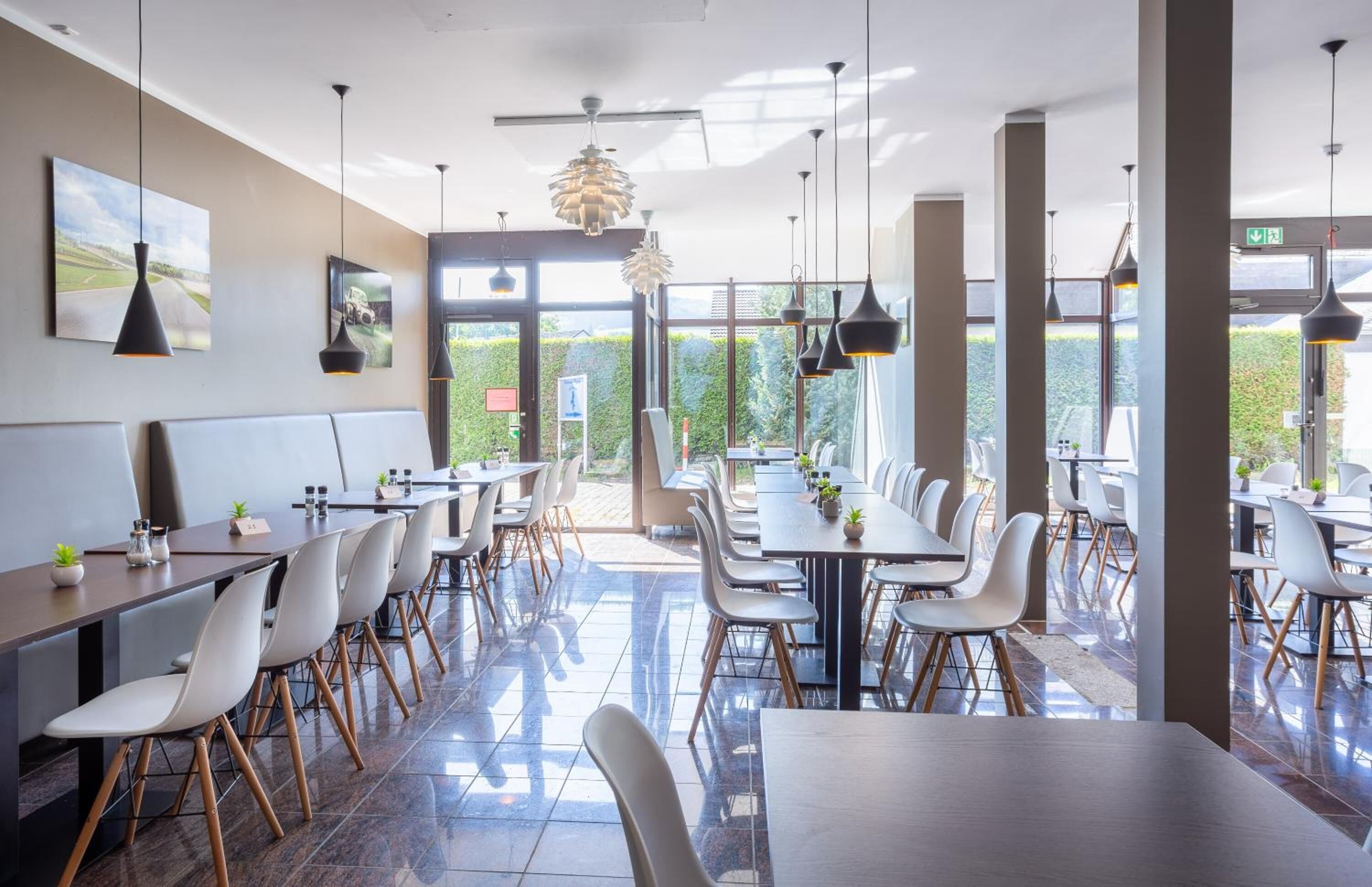 A modern dining area with long tables and white chairs, large windows with a view of greenery, and pendant lighting.