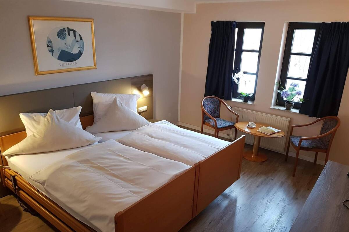 Une chambre d’hôtel avec un grand lit, deux chaises, une petite table et une fenêtre avec des rideaux.