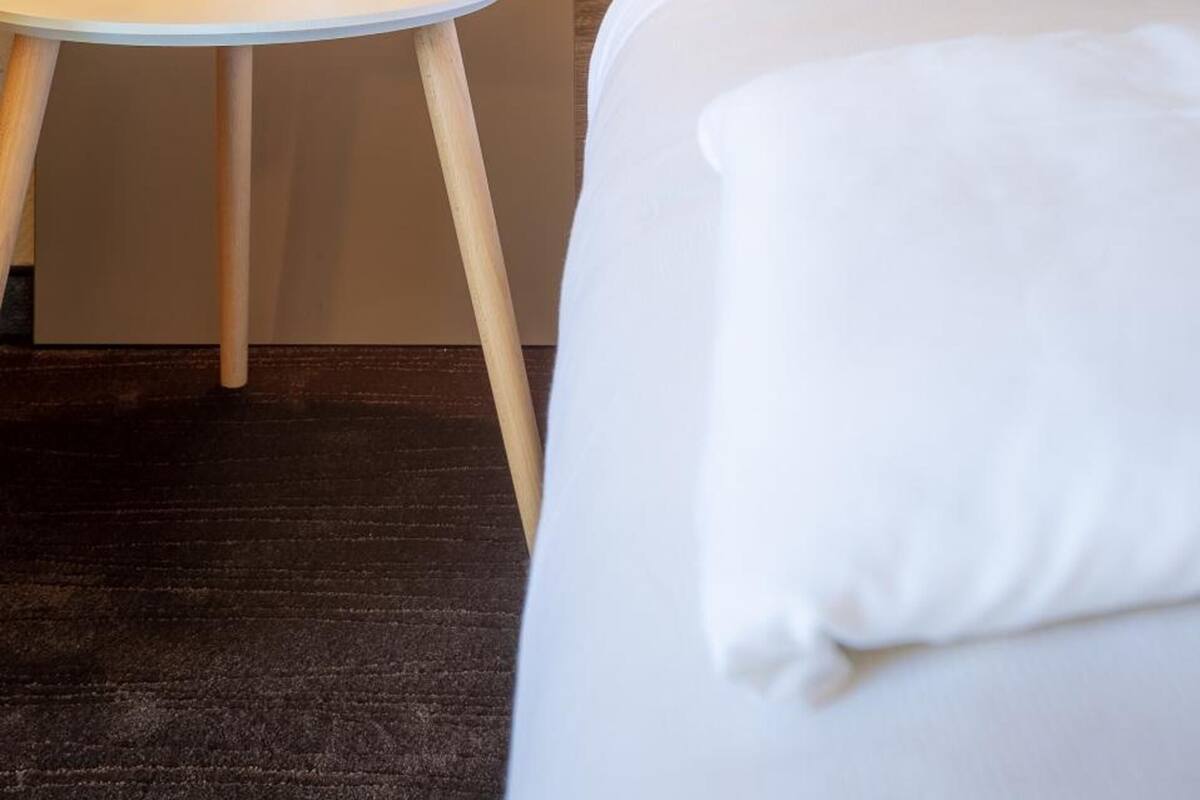 Une chambre d’hôtel avec un lit, une petite table ronde sur laquelle se trouvent une tasse et une plante, et un luminaire fixé au mur.