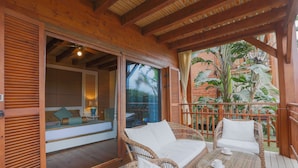 Una terraza de madera con mobiliario de mimbre, una puerta de cristal que da a una habitación y vistas a plantas tropicales.