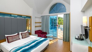 Habitación de hotel con una cama grande, un sofá rojo, una mesita, un televisor y un balcón con vistas a palmeras.