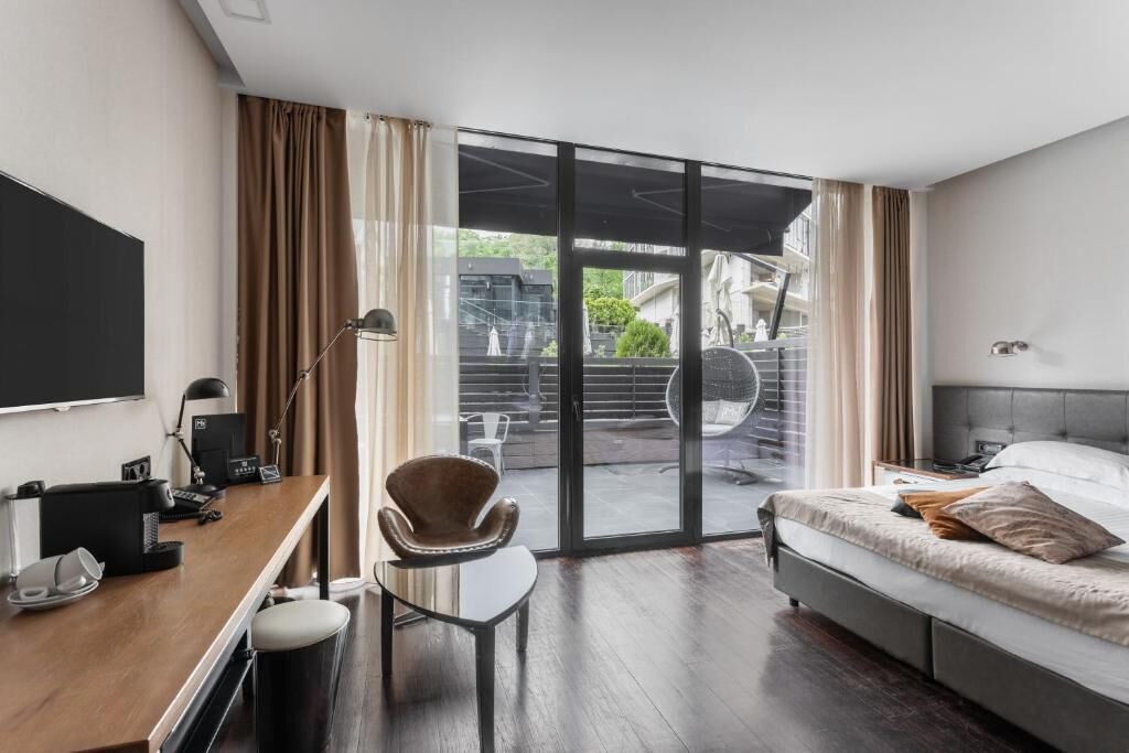 A modern hotel room with a bed, desk, chair, and a large sliding glass door leading to an outdoor area.