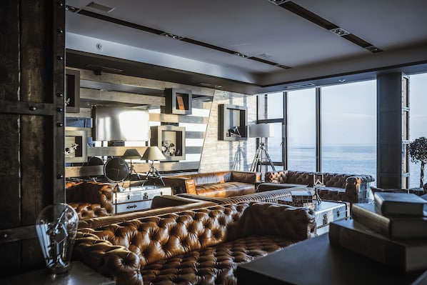 A modern living room with a large window overlooking the sea, featuring leather sofas, a coffee table, and a bookshelf.