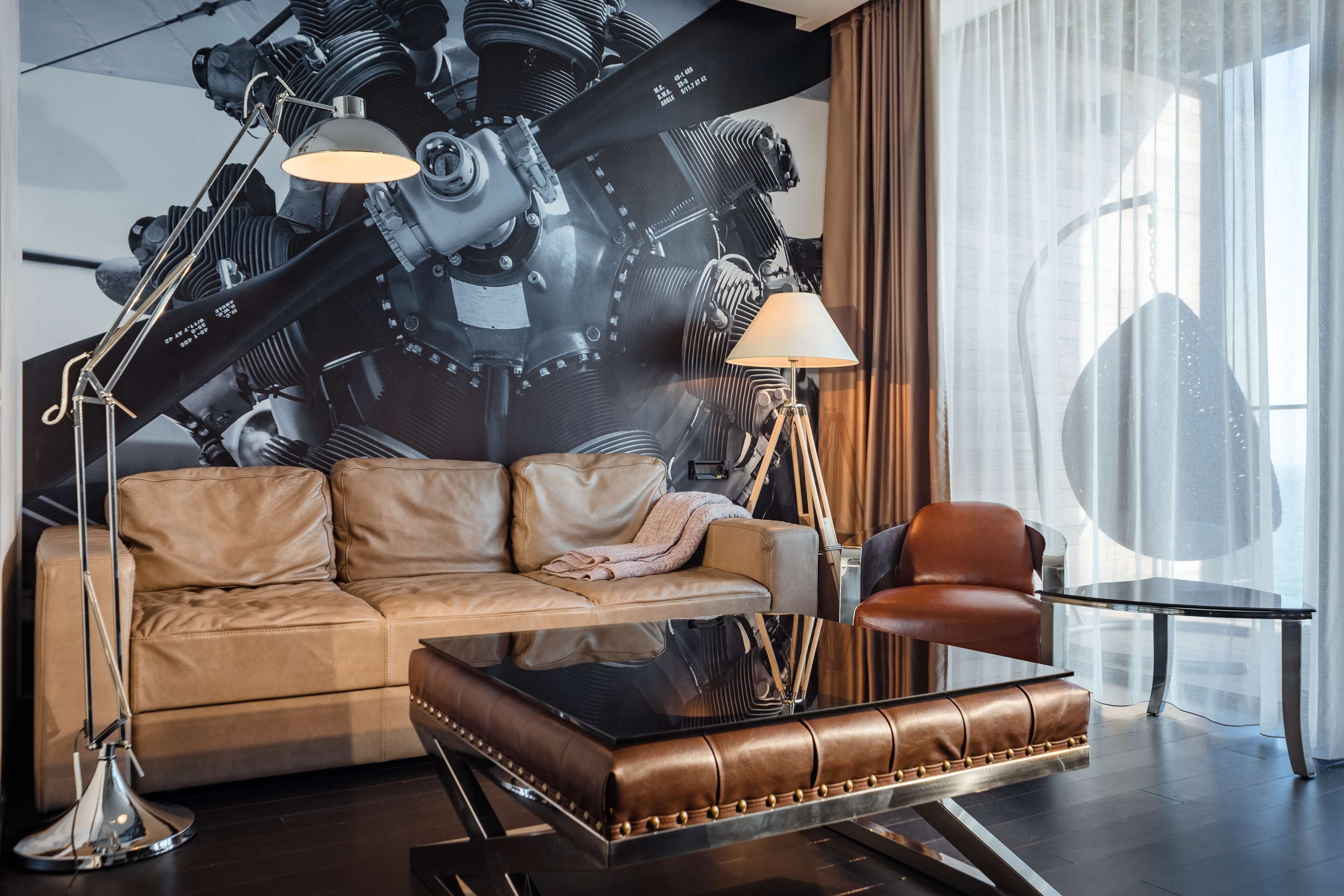 Uma sala de estar moderna com um sofá de couro, um banquinho de couro marrom, uma mesa de centro de vidro, um abajur e um grande quadro abstrato na parede.