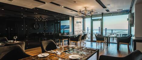 Un restaurante moderno con vistas al mar, que combina decoración oscura y clara, y ofrece diferentes opciones de asientos.