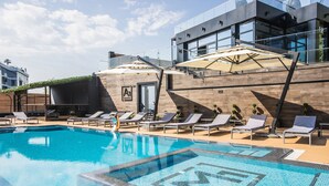 Una piscina con lettini e ombrelloni, circondata da un edificio moderno con ampie finestre e facciata in legno.