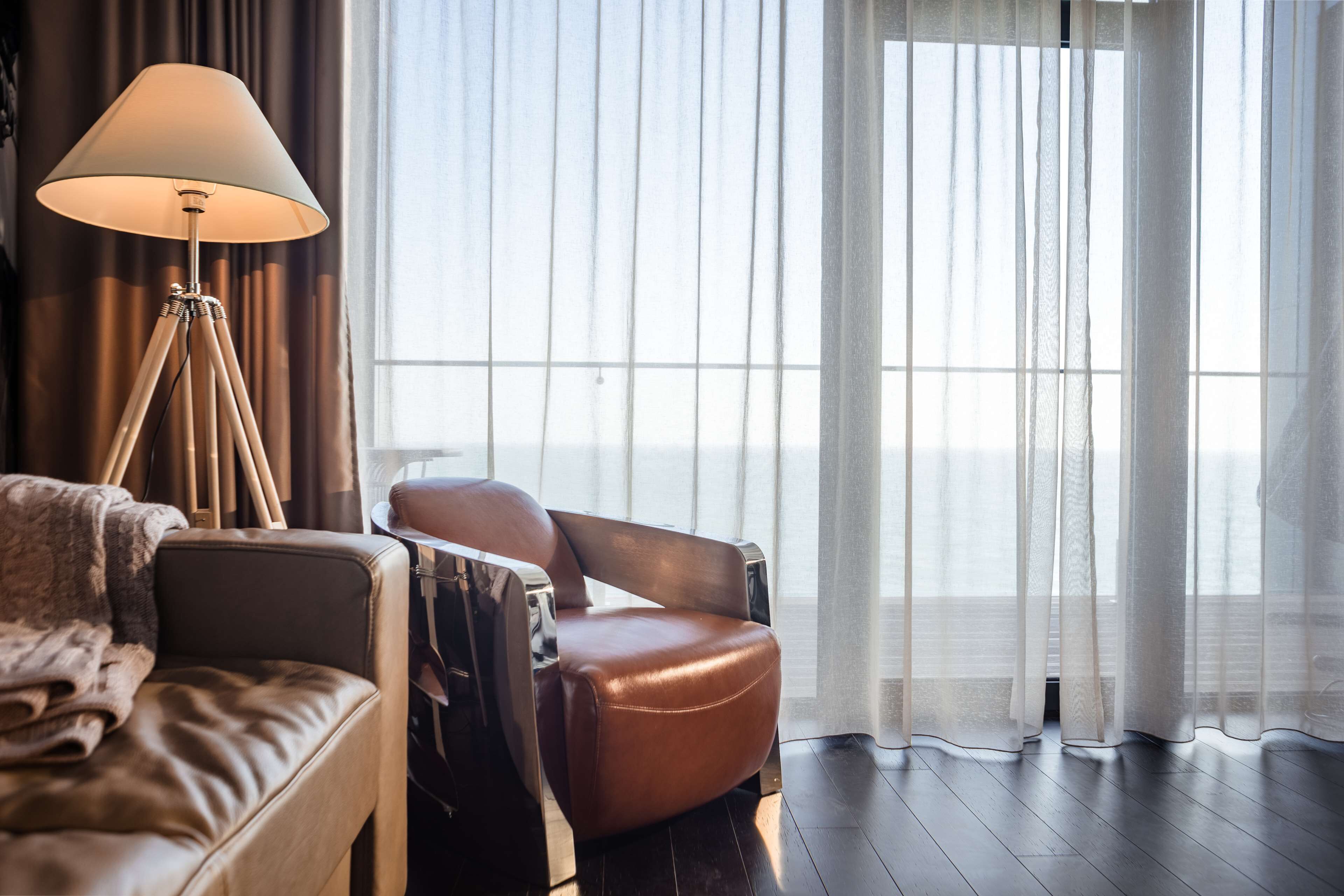 A modern hotel room with a leather sofa, a reclining chair, a floor lamp, and a large window with sheer curtains.