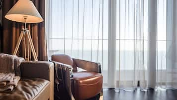 A modern hotel room with a leather sofa, a reclining chair, a floor lamp, and a large window with sheer curtains.