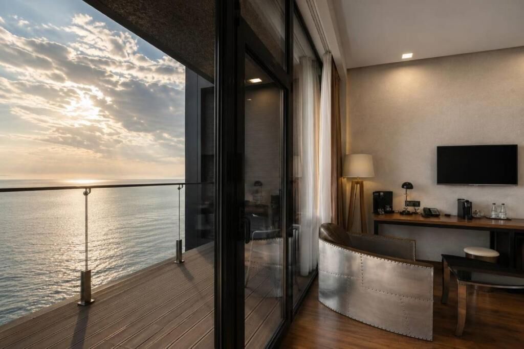 Un balcón con vista al mar, una zona de estar con sofá y sillones, un televisor de pantalla plana y un escritorio con lámpara.