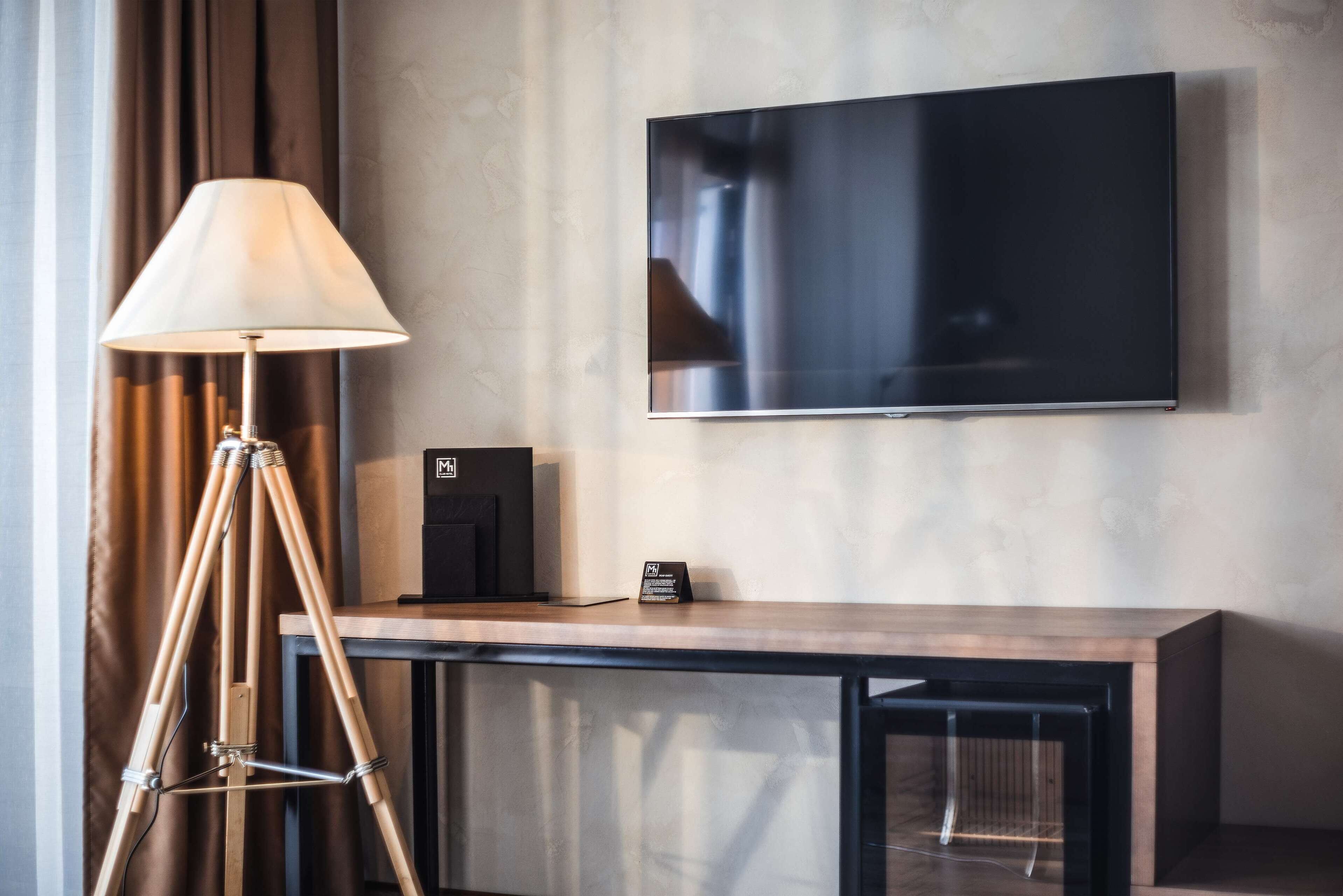 Quarto de hotel moderno com uma grande televisão de tela plana na parede, uma escrivaninha de madeira com uma luminária e um pequeno dispositivo preto sobre a escrivaninha.