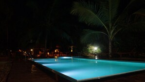 A swimming pool at night with palm trees and a building in the background. - Athgira River Camp (Udawalawa)