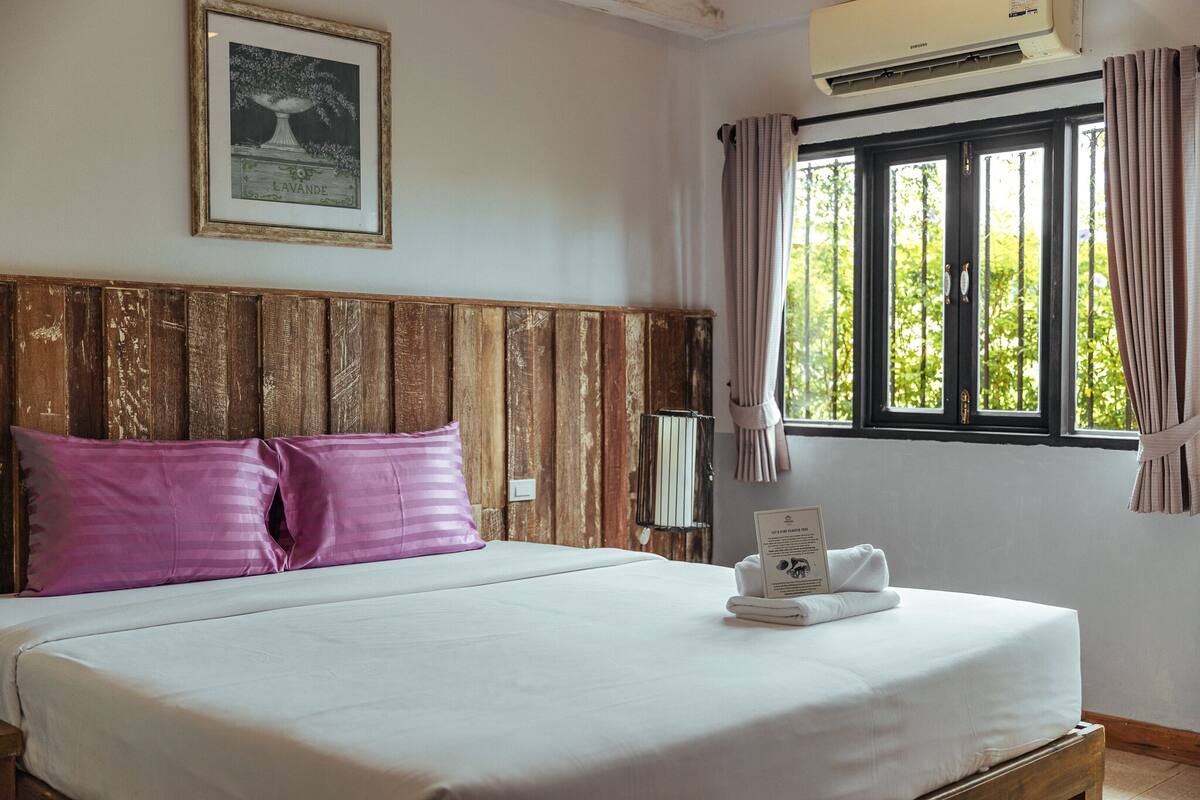 A neatly made bed with white linens and two pink pillows, a framed picture on the wall, and a window with curtains.