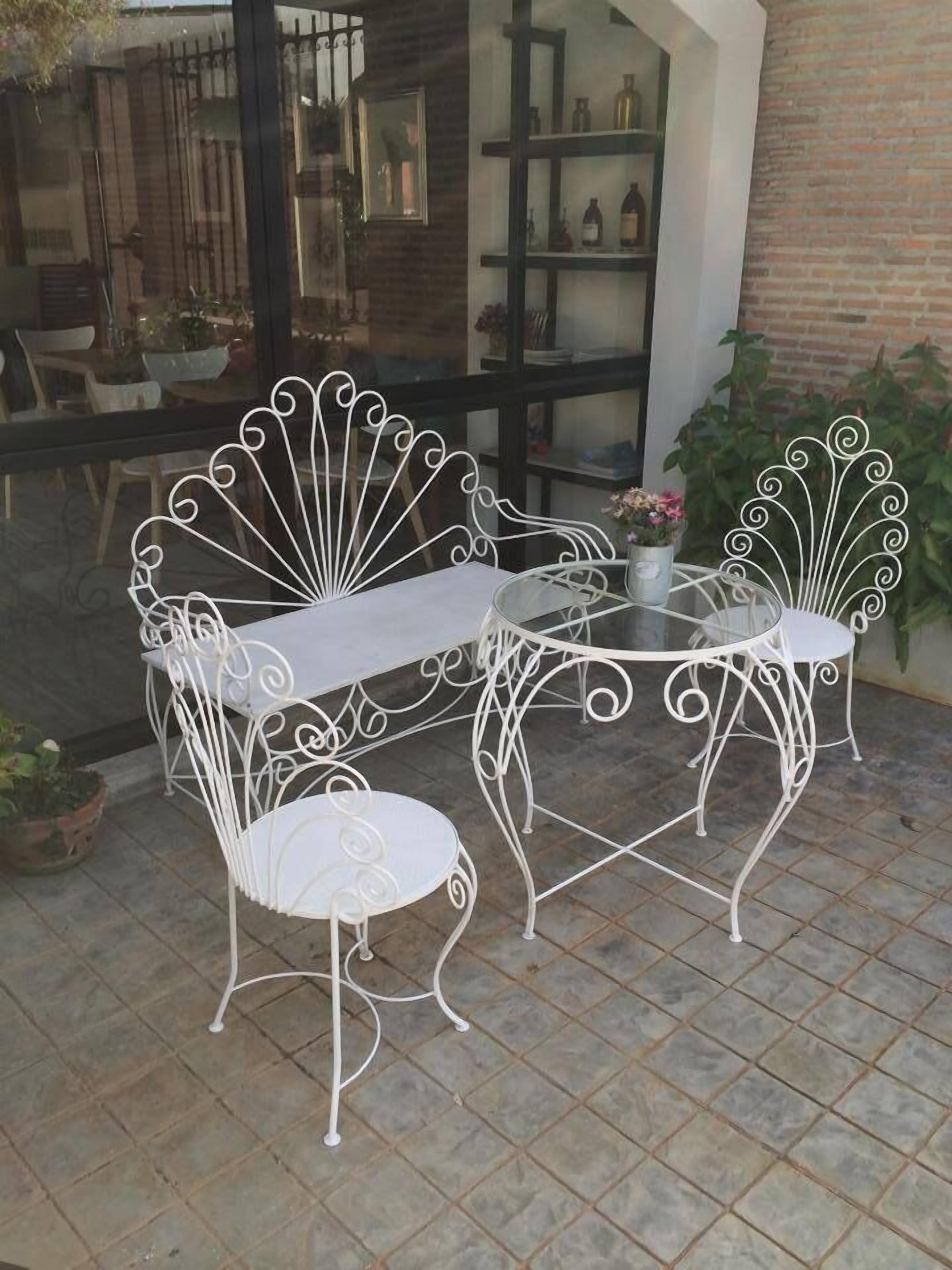A white metal outdoor dining set with a glass table and chairs, positioned on a tiled patio with a brick wall and potted plants in the background.
