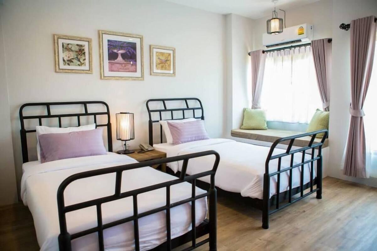 A bedroom with two beds, a bedside table with a lamp, a window with curtains, and framed pictures on the wall.