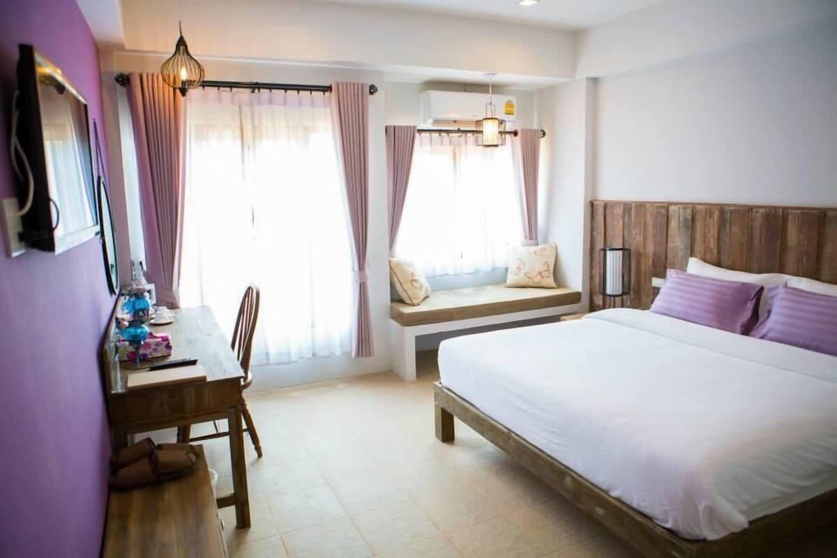 A bedroom with a wooden bed, a bench with pillows, a desk with a chair, and a window with curtains.