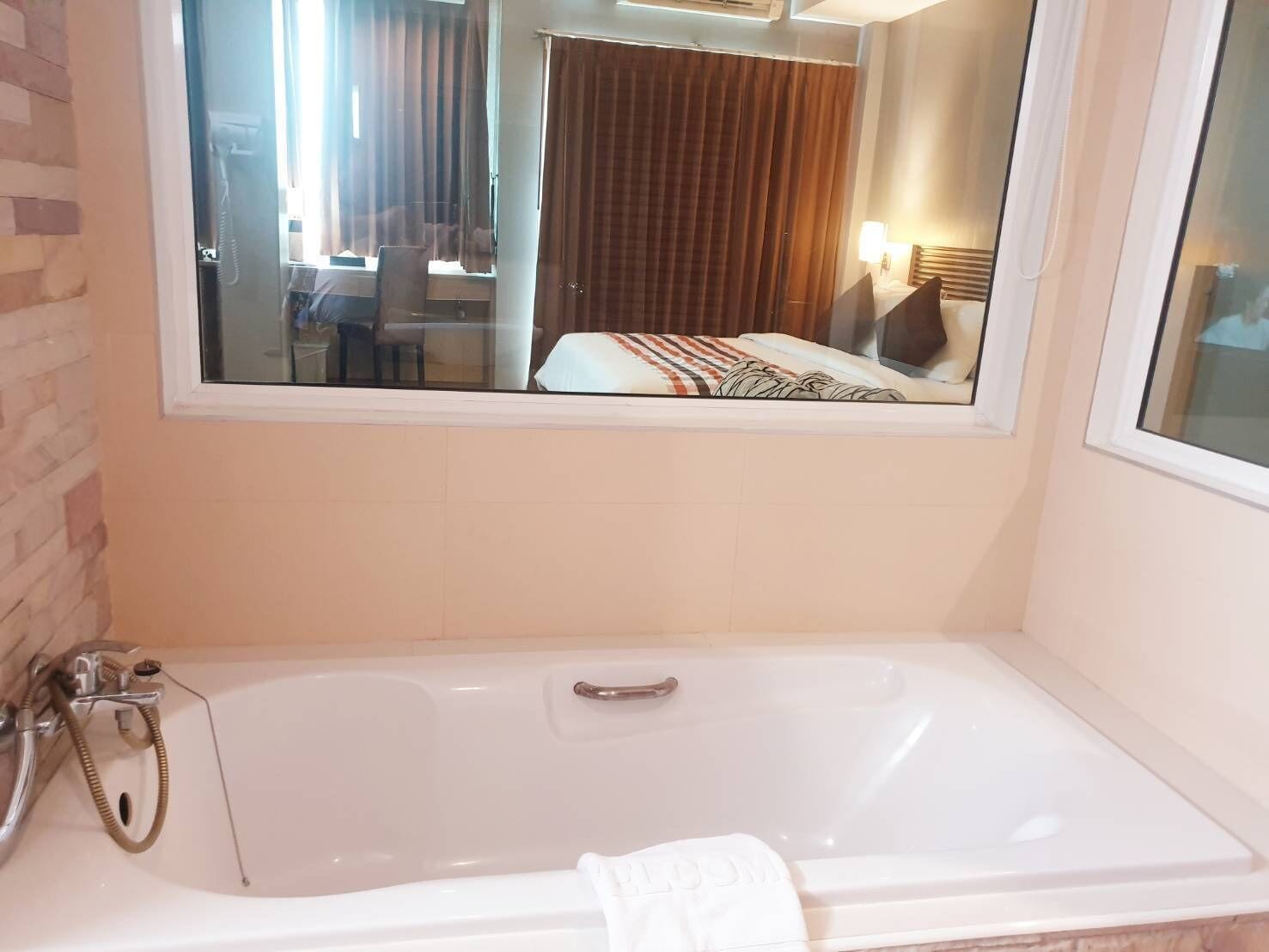 A bathroom with a bathtub, a shower, and a mirror reflecting a room with a bed and curtains.