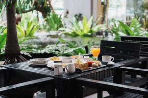 Una mesa dispuesta para el desayuno, con tostadas, huevos y jugo, mostrando una vista de vegetación exuberante y una fuente de agua al fondo.