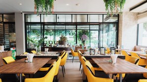 A restaurant with yellow chairs and wooden tables, large windows, and hanging plants. - Green Hotel & Resort Khon Kaen (Khon Kaen)