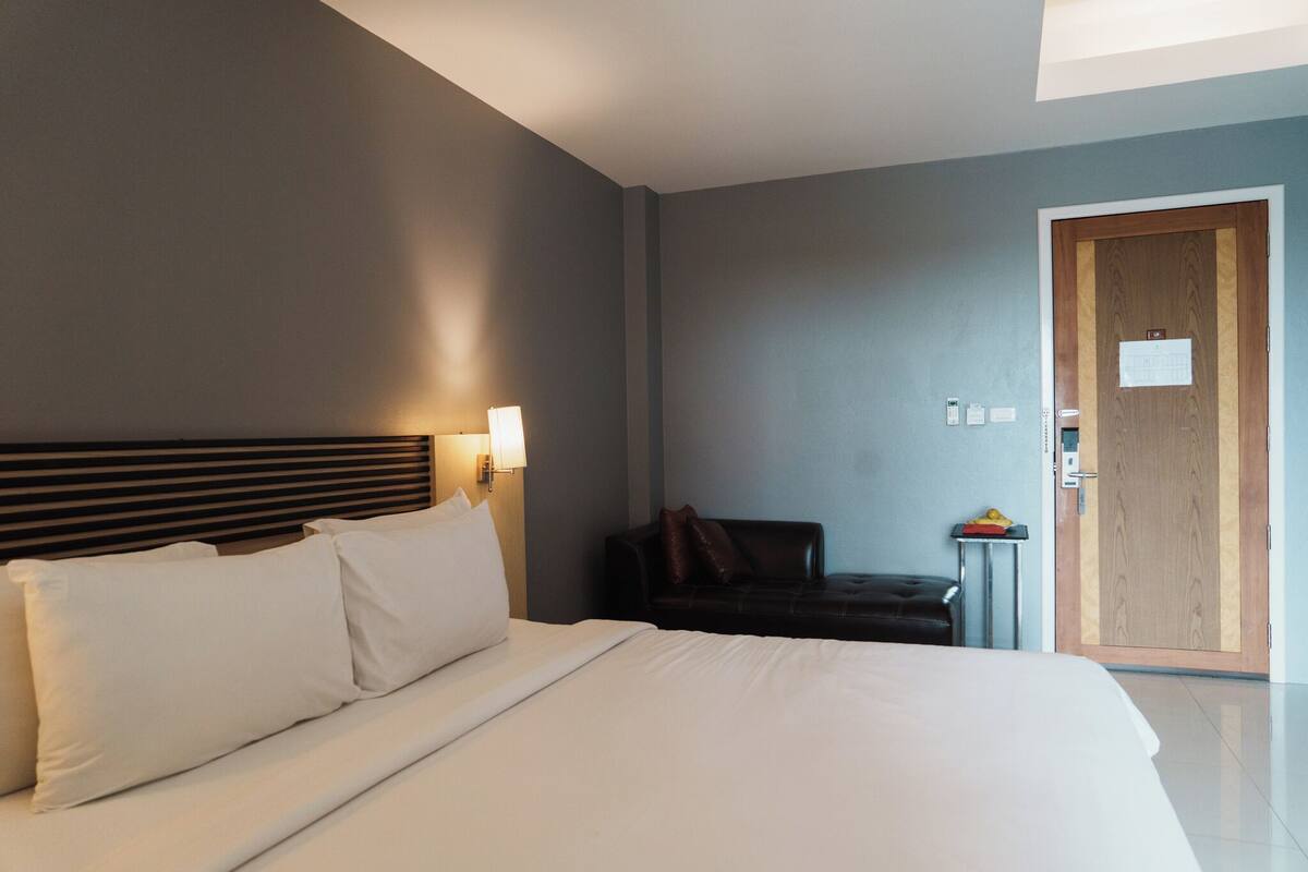 A hotel room with a large bed, a wooden headboard, a small table with a lamp, a dark leather chair, and a wooden door.