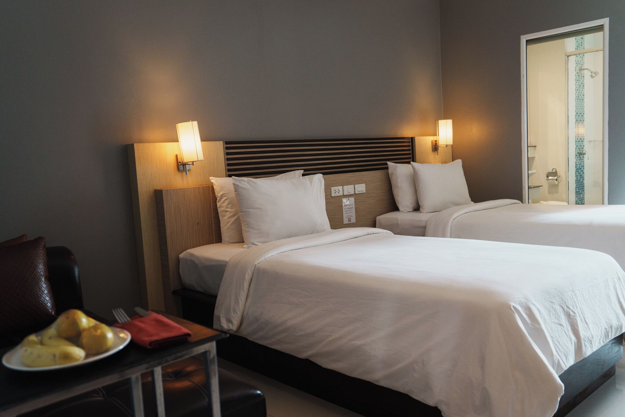Une chambre d’hôtel avec deux lits, une table de chevet sur laquelle se trouve une assiette de fruits et des lampes fixées au mur.