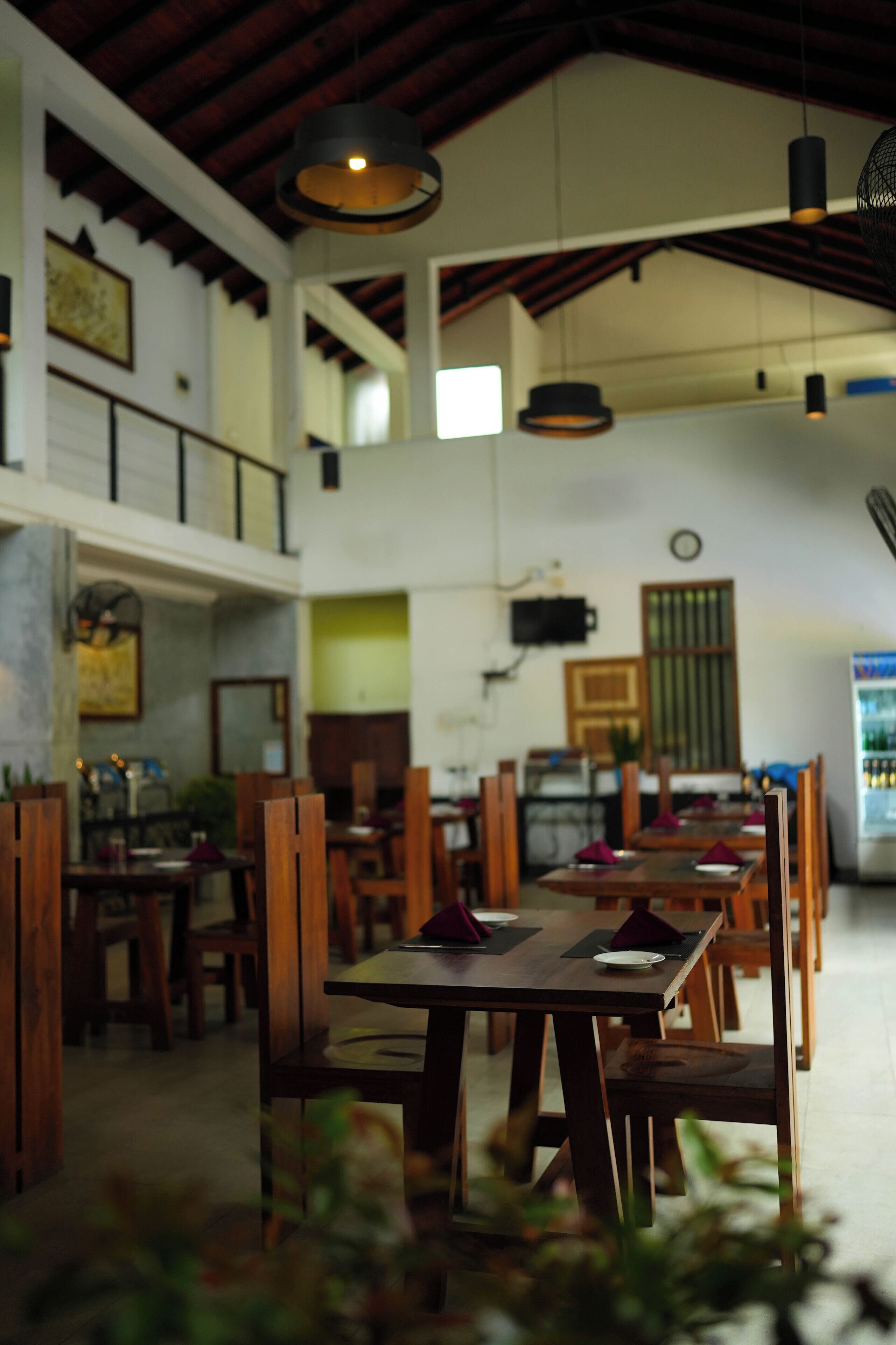A restaurant with wooden tables and chairs, hanging pendant lights, and a high ceiling.