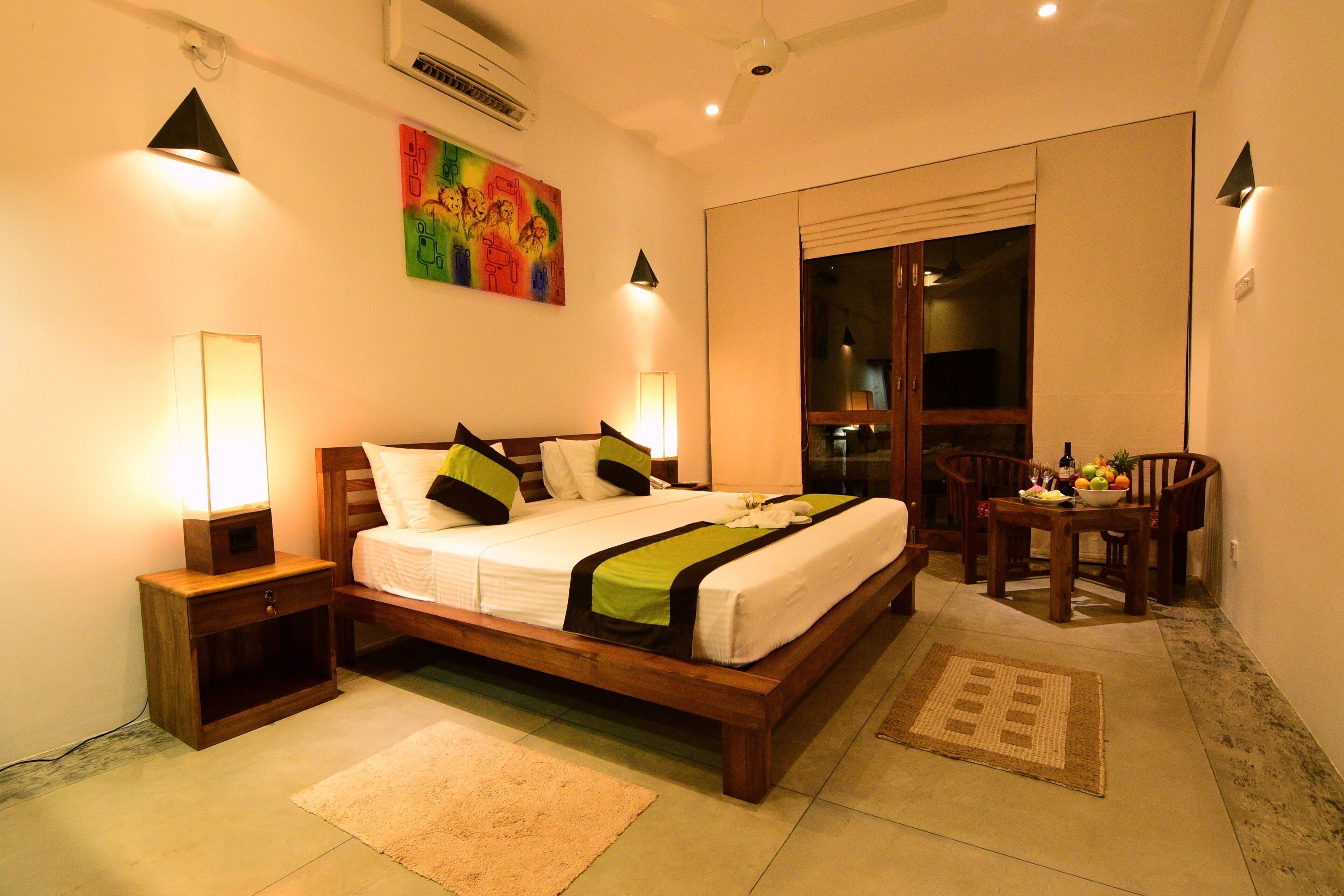 A well-lit bedroom with a wooden bed, bedside tables, a colorful artwork on the wall, and a dining area with chairs and a table.