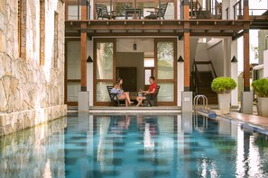 A pool area with a stone wall, a wooden deck with chairs, and a staircase leading to an upper level.