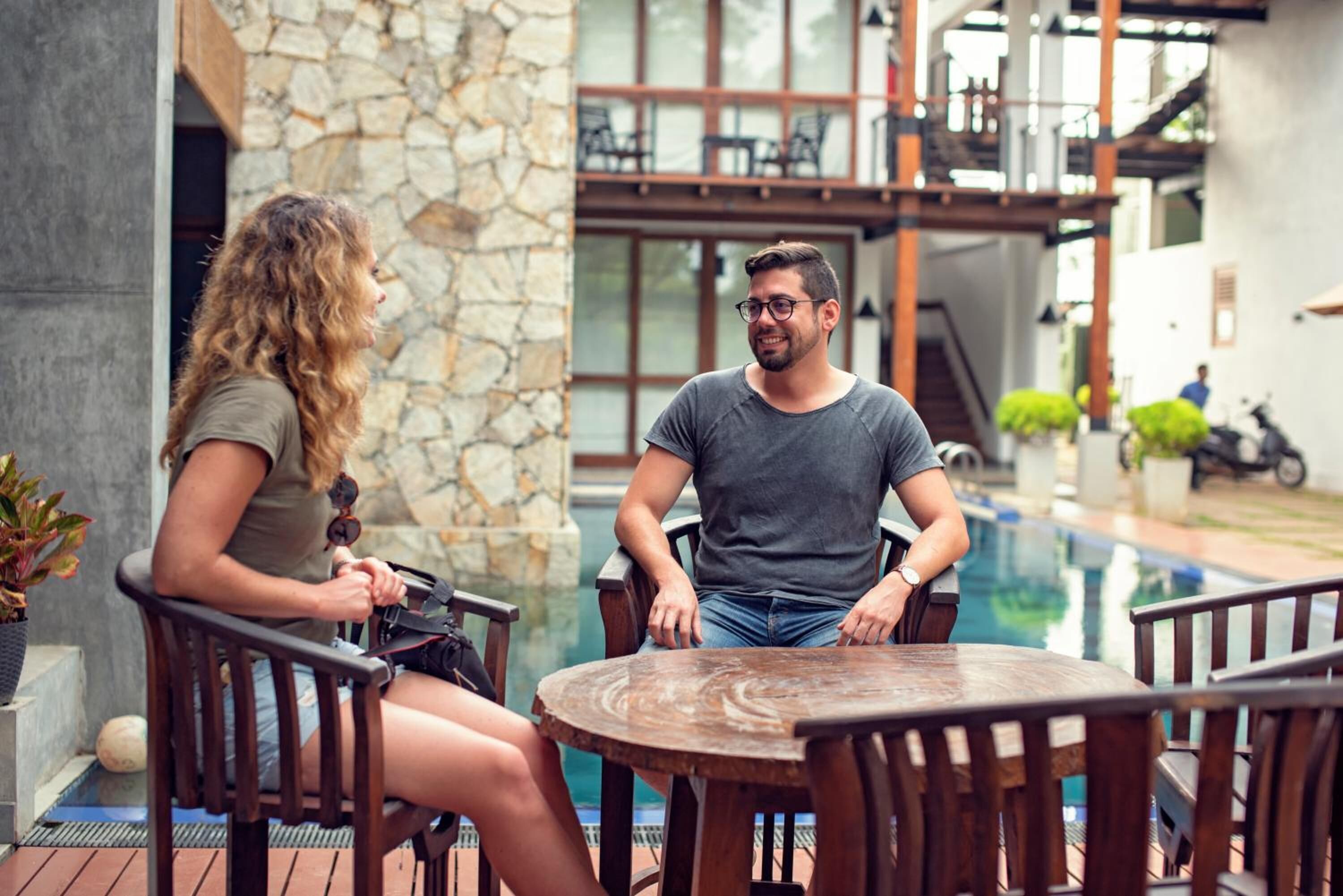 A couple sitting at a wooden table by a poolside, with stone walls and a modern interior in the background.