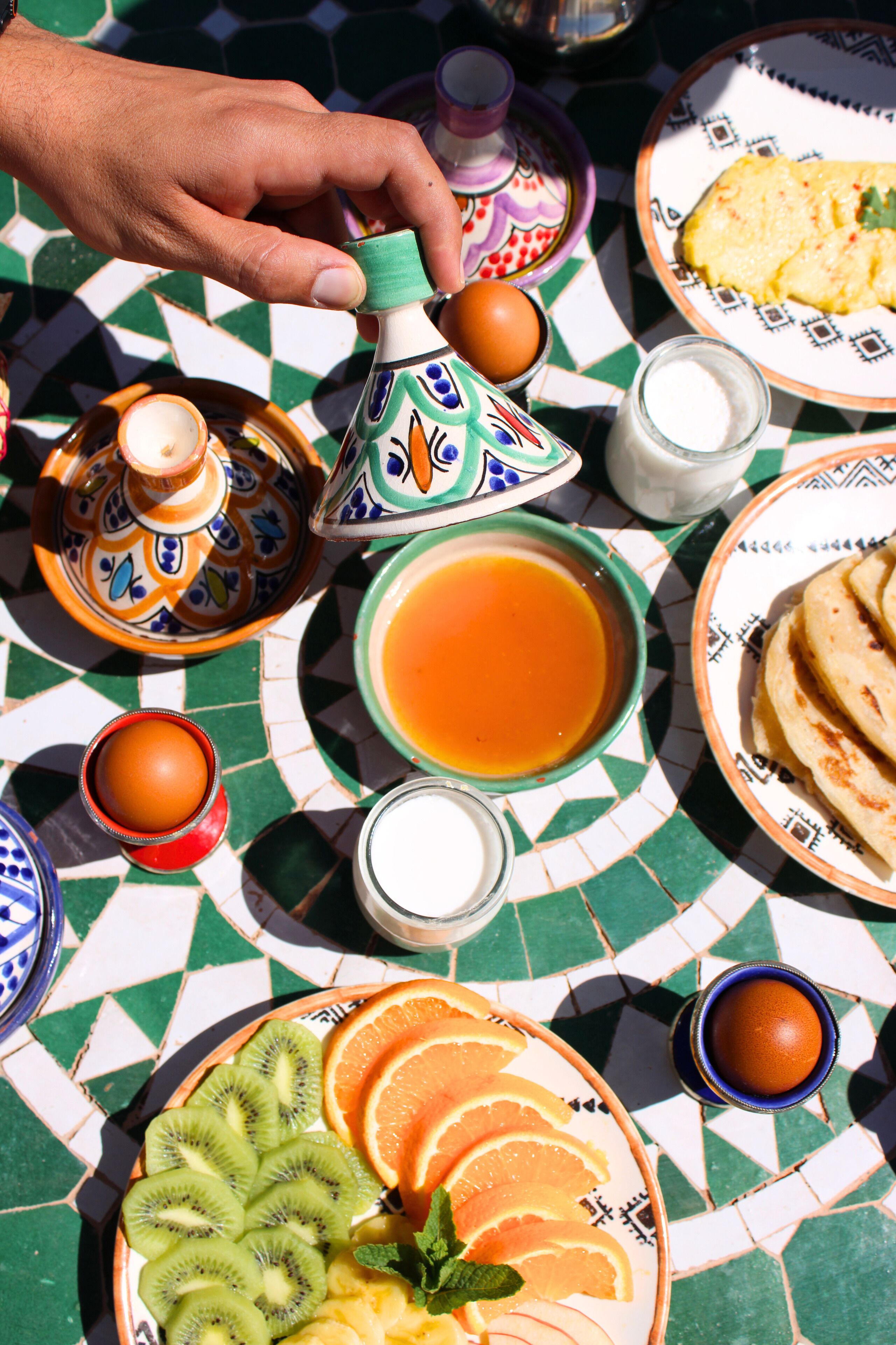 A table with a Moroccan setting, including a teapot, plates with food, and a hand reaching for a teapot.