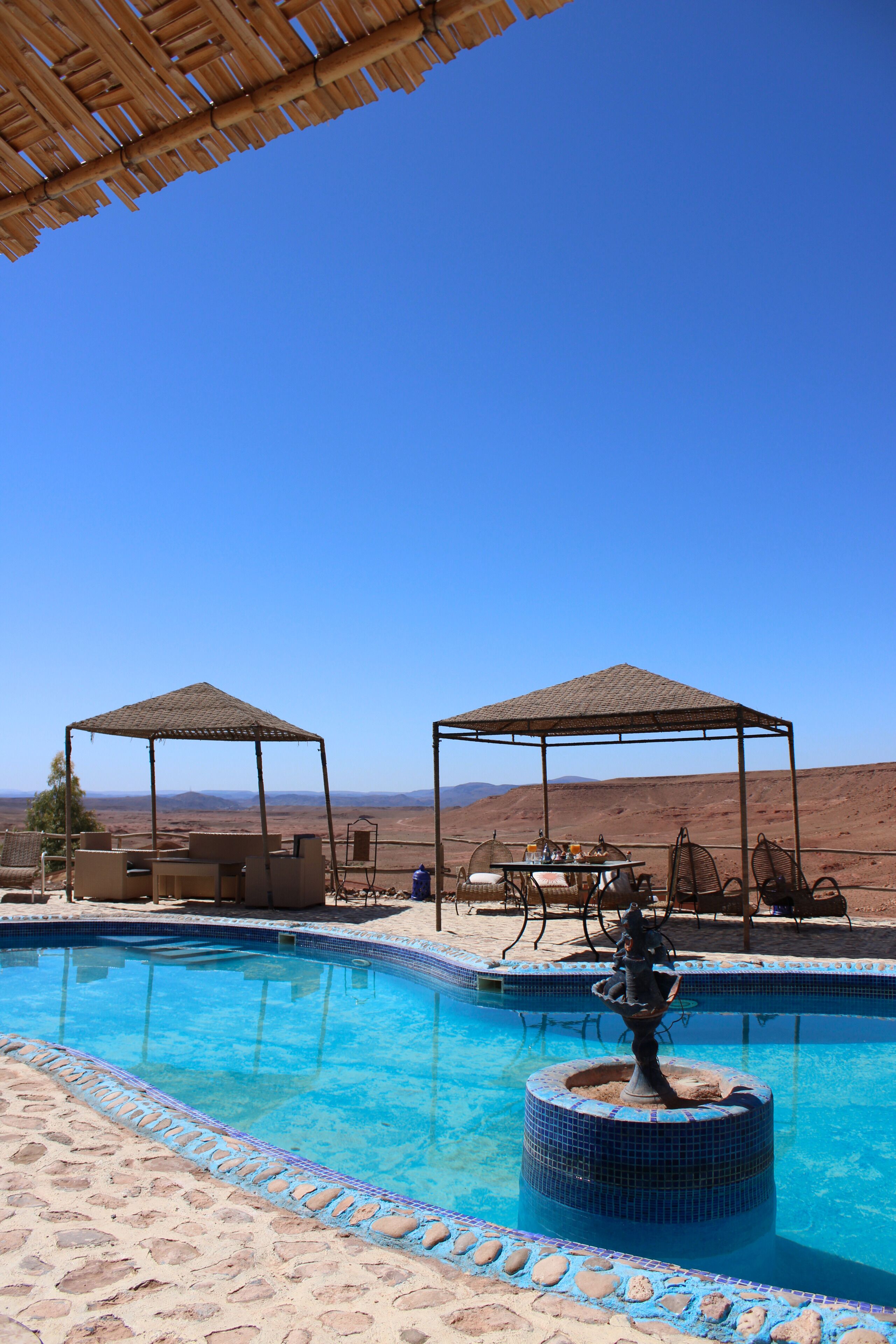 A pool with a fountain, two shaded seating areas, and a view of the desert landscape.