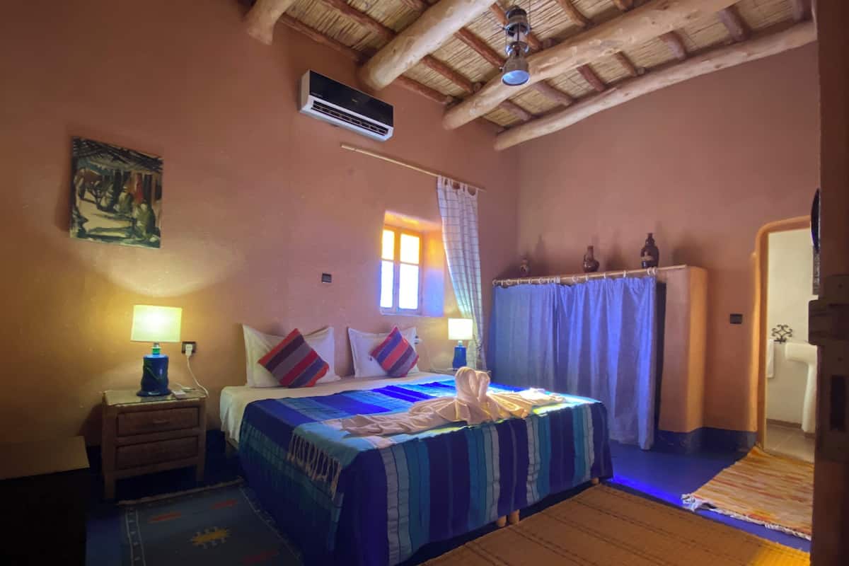 A bedroom with a bed, bedside table, lamp, air conditioning unit, and a window with curtains.