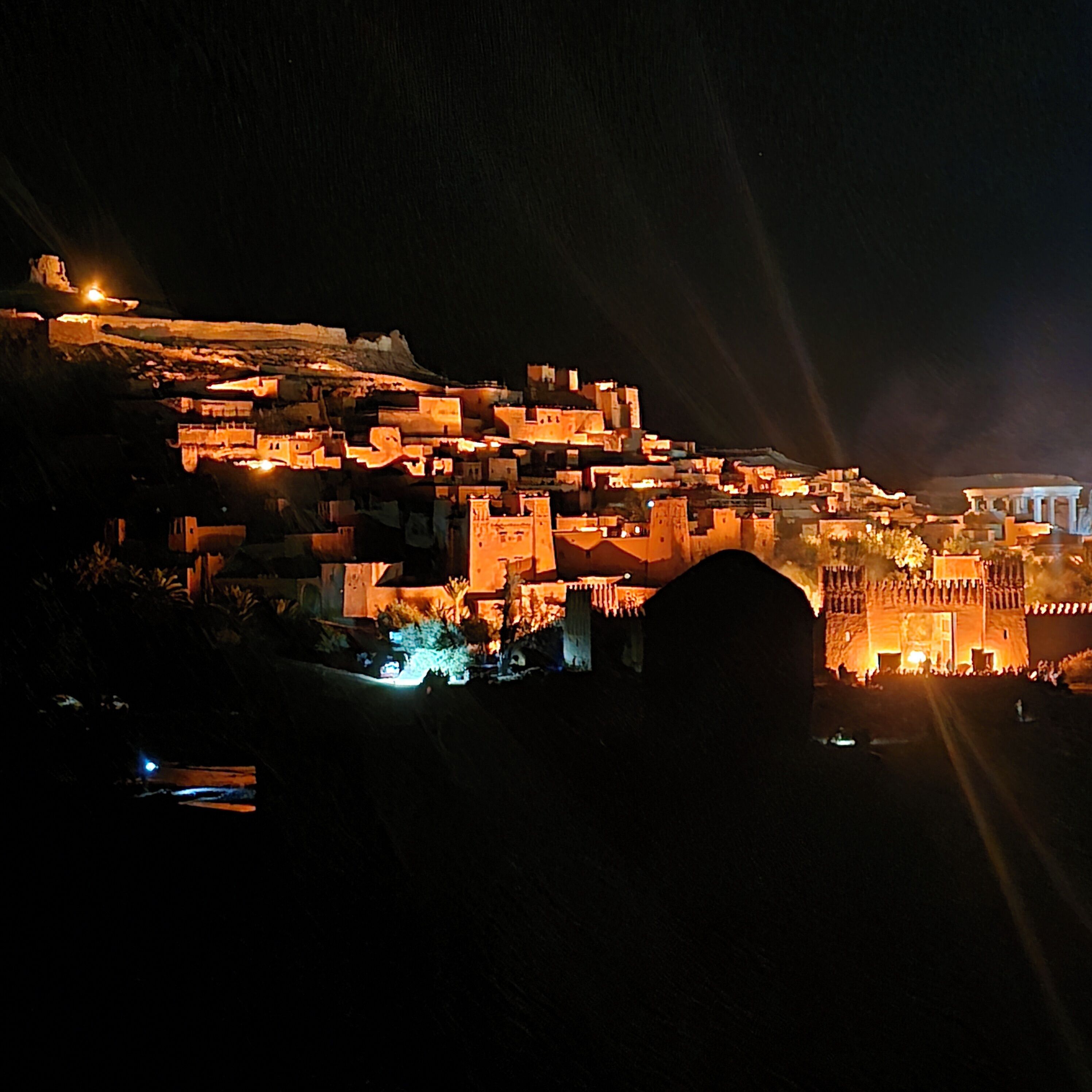 Vista nocturna de una ciudad amurallada histórica, con edificios iluminados y una estructura antigua destacada.