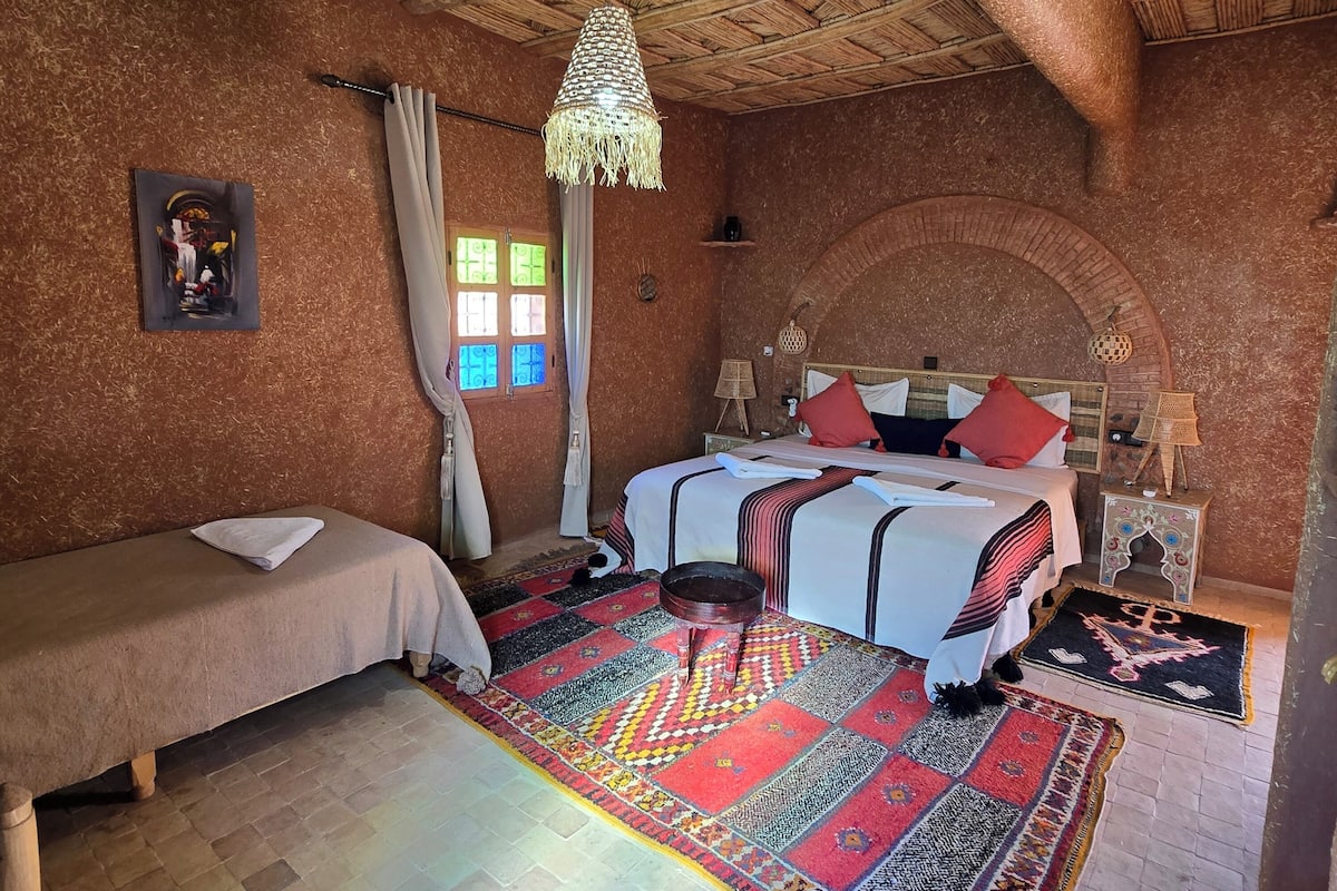 A room with a thatched ceiling, a bed with a colorful rug, a small table with a lamp, and a window with curtains.