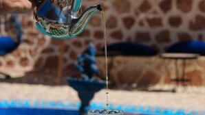 Una mano vertiendo té desde una tetera de metal en un vaso, con azulejos de estilo marroquí, una pared de piedra y una piscina al fondo.