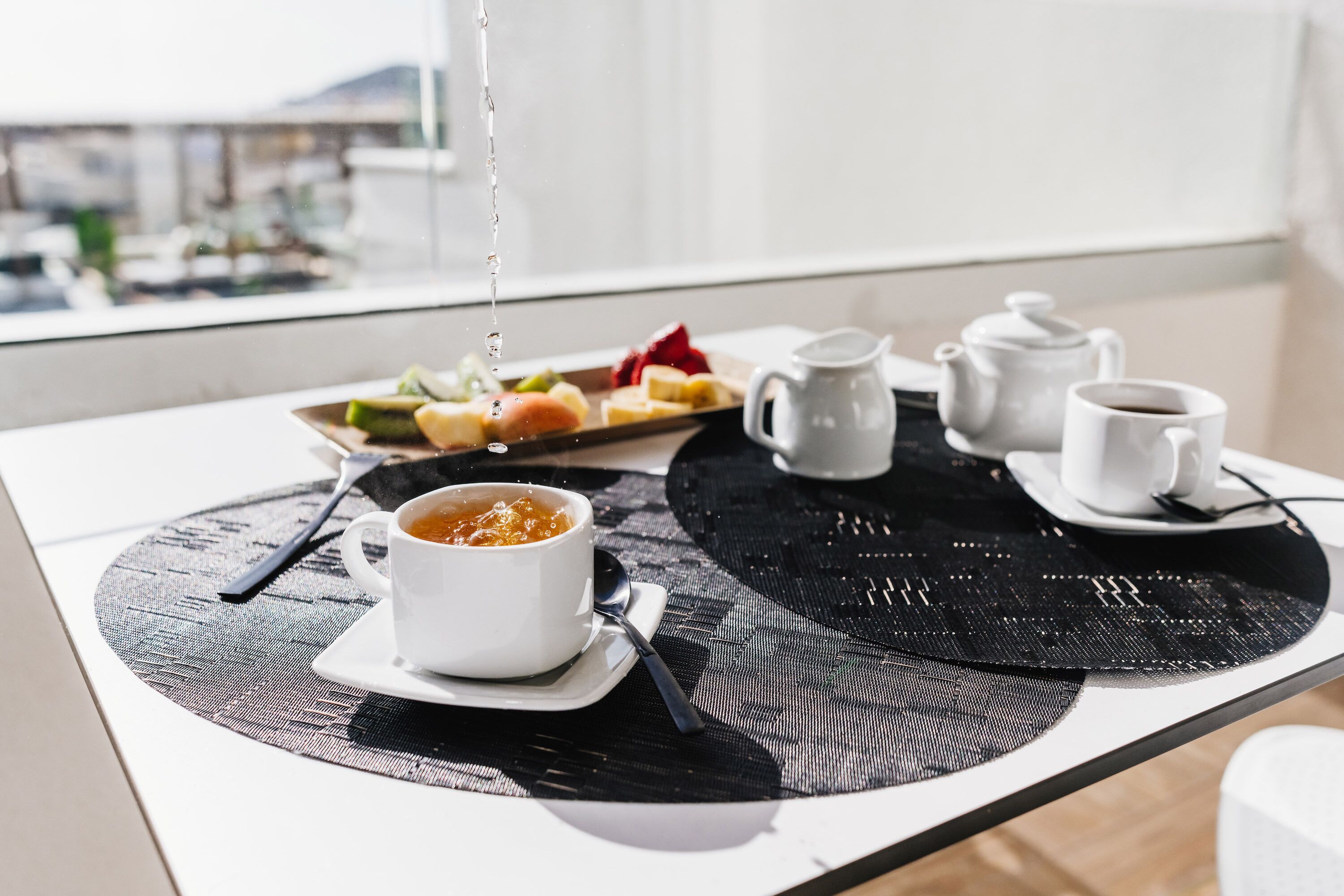 A breakfast setup with a bowl of soup, a cup of coffee, and a plate of fruit on a table with a black placemat.