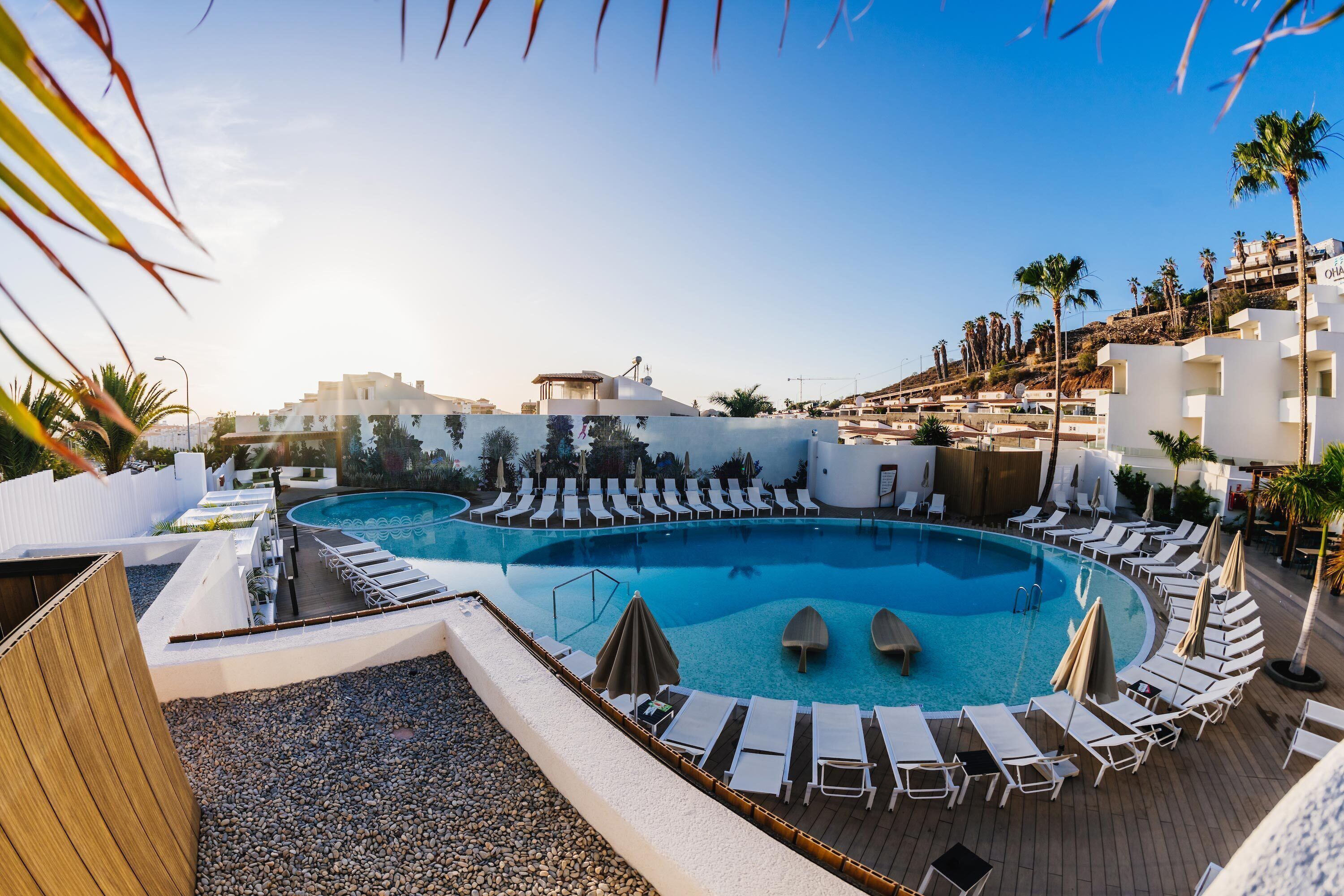 Un área de piscina con sillones de descanso y sombrillas, rodeada de edificios y palmeras.