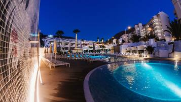 Zona de piscina con sillas de descanso y una piscina, rodeada de edificios y palmeras.