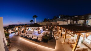 Un espace de détente extérieur moderne, doté d’un plancher en bois, de différents coins salon et d’une piscine en arrière-plan.