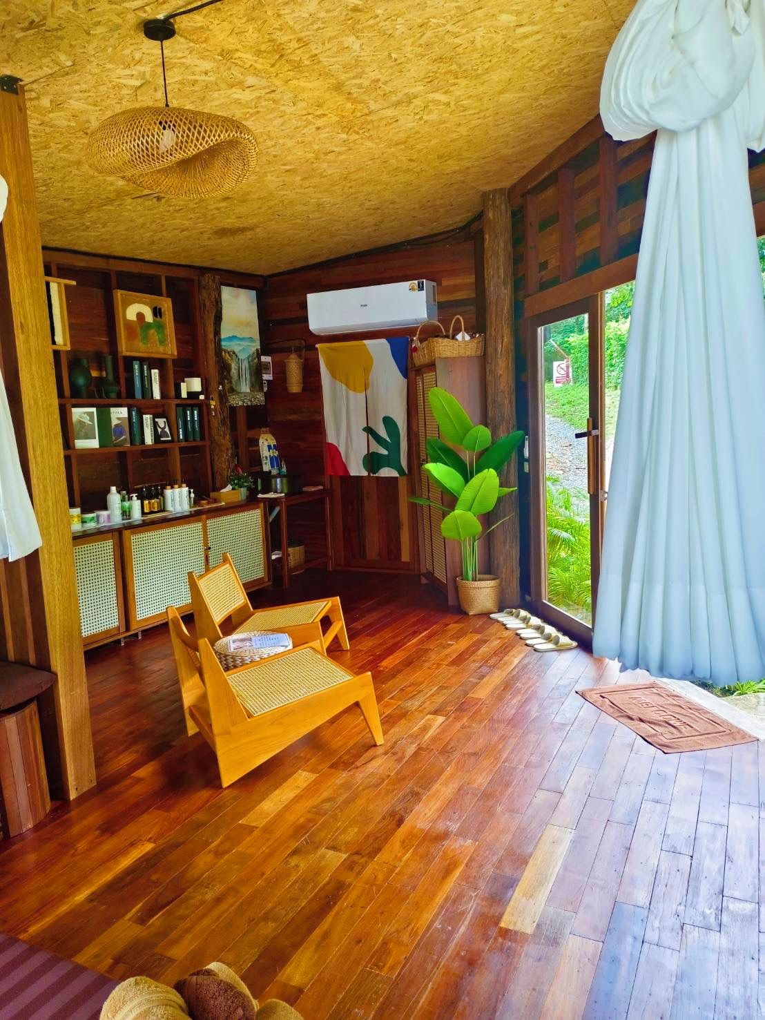 Una habitación con piso de madera, una zona de estar con sillas, una estantería, una lámpara colgante y vistas a través de una puerta de cristal.