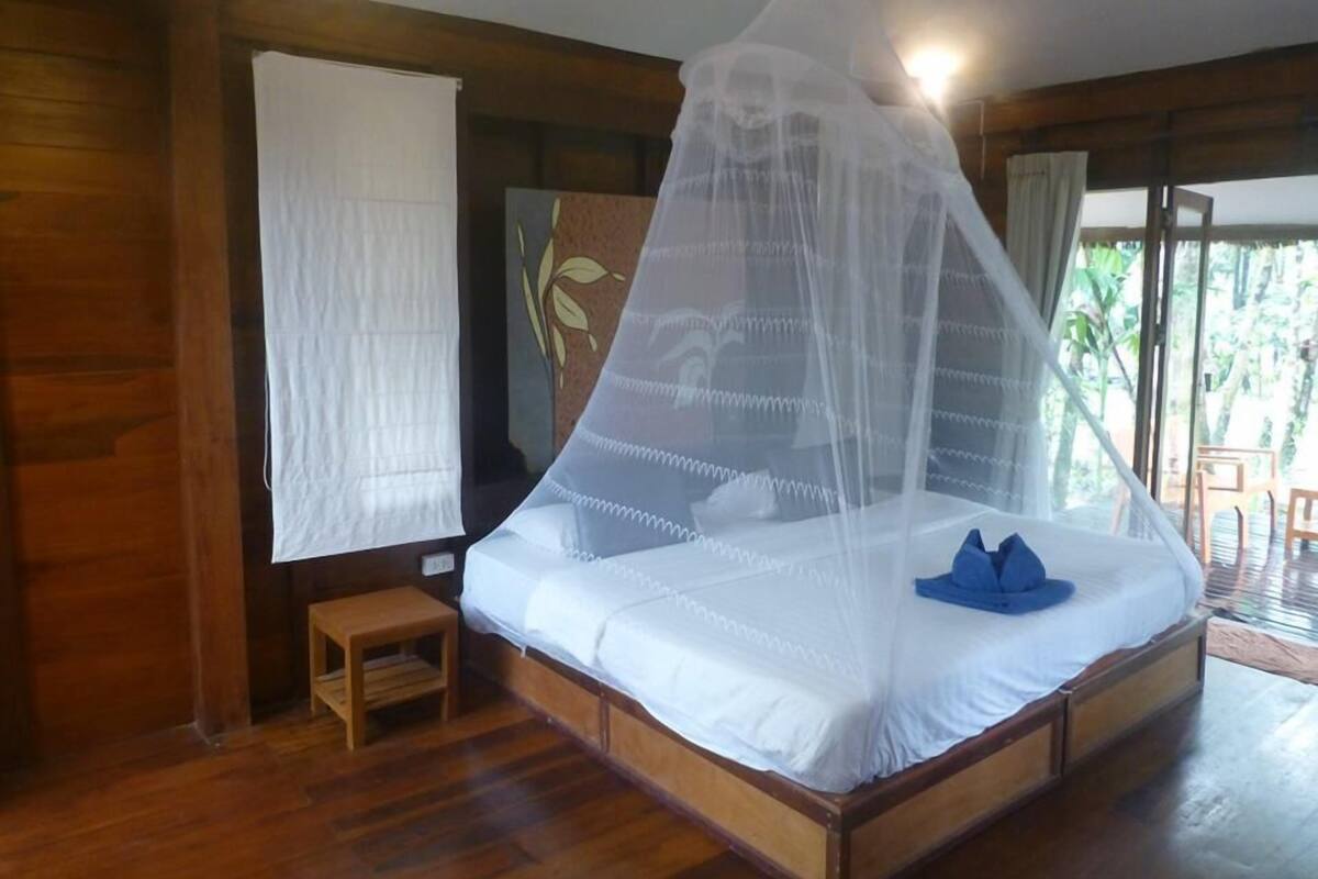 A wooden-framed bed with a white mattress and a mosquito net. There is a small wooden stool beside the bed and a window with a white curtain.